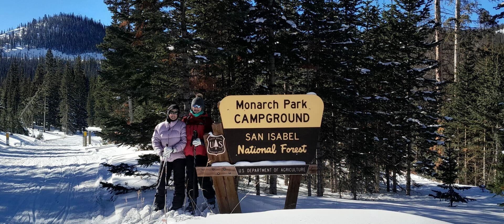 Winter day, blue sky, snow all around in the forest with 2 ladies cross country skiing standing by a sign that says Monarch Park.