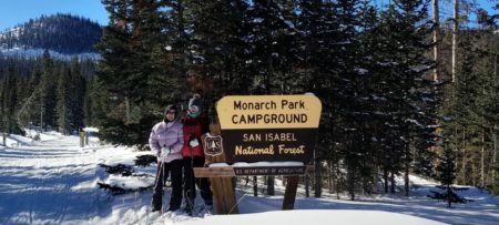 Winter day, blue sky, snow all around in the forest with 2 ladies cross country skiing standing by a sign that says Monarch Park.