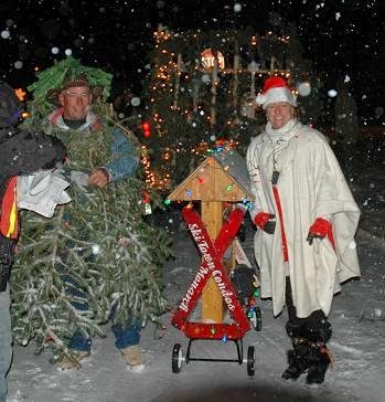 Winter snowy evening at a parade with a man dressed up as a Christmas tree and a lady dressed up as Mrs. Claus standing next to a mini Ski Town Condos float with Christmas lights shining.