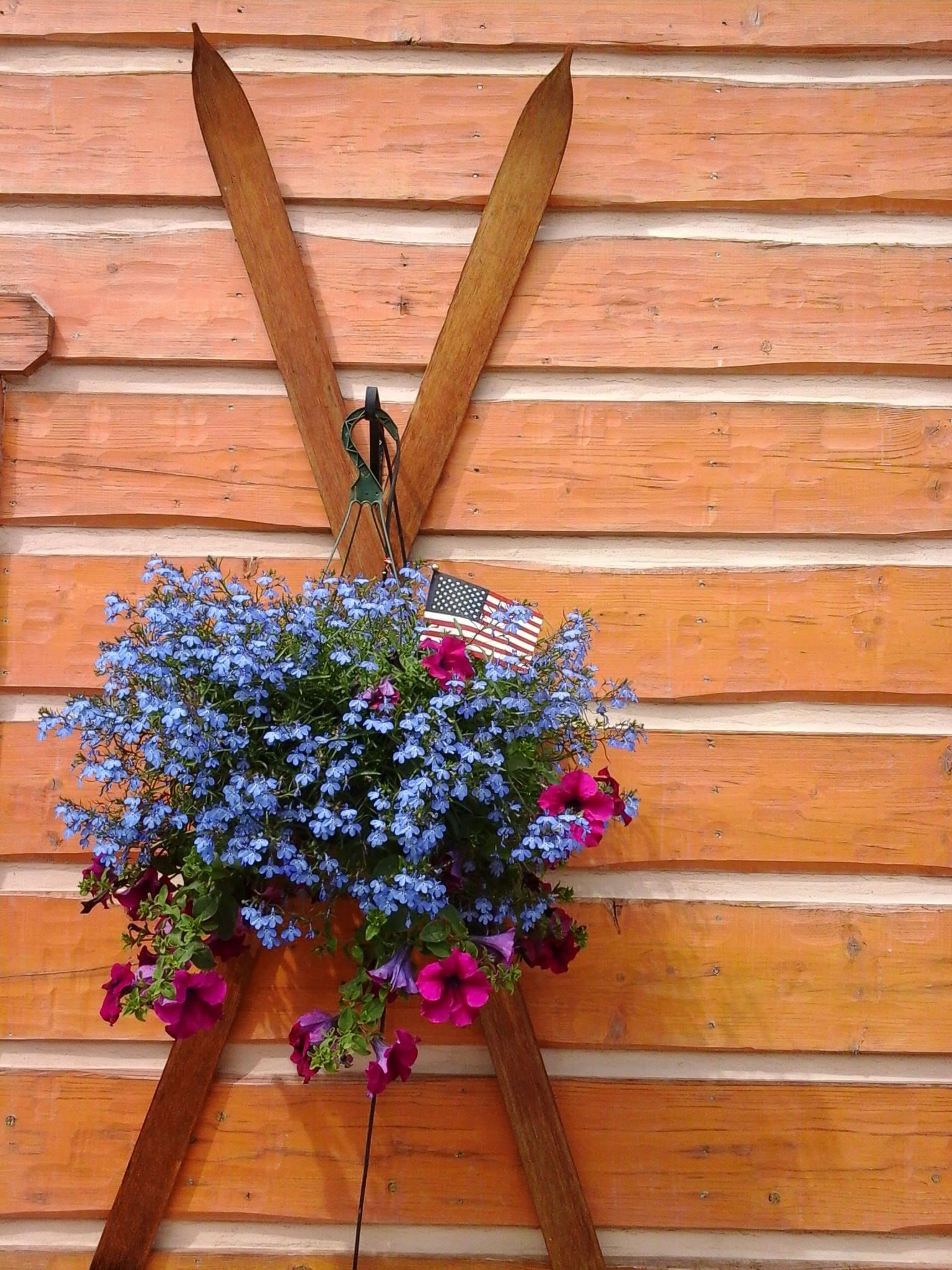 Exterior photo of a large hanging flower basket with purple and pink flowers and an American flag.  Basket hanging over antique skies on  log sided condo.