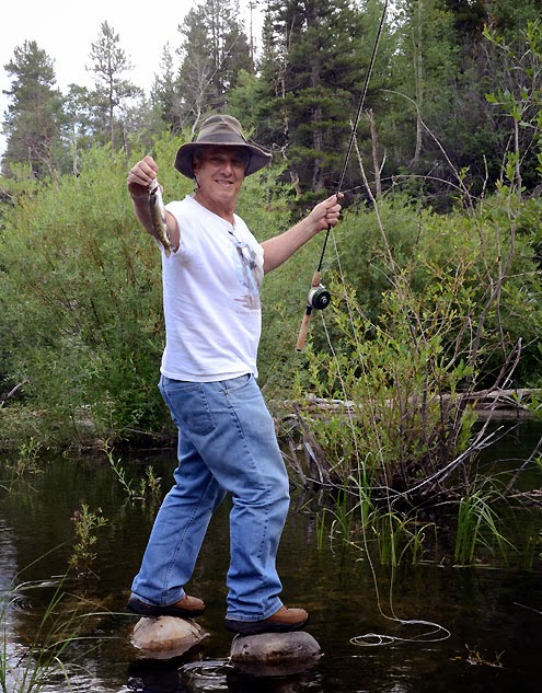 Man standing on two rocks smiling with a fish on the hook of his fishing pole in the wilderness.