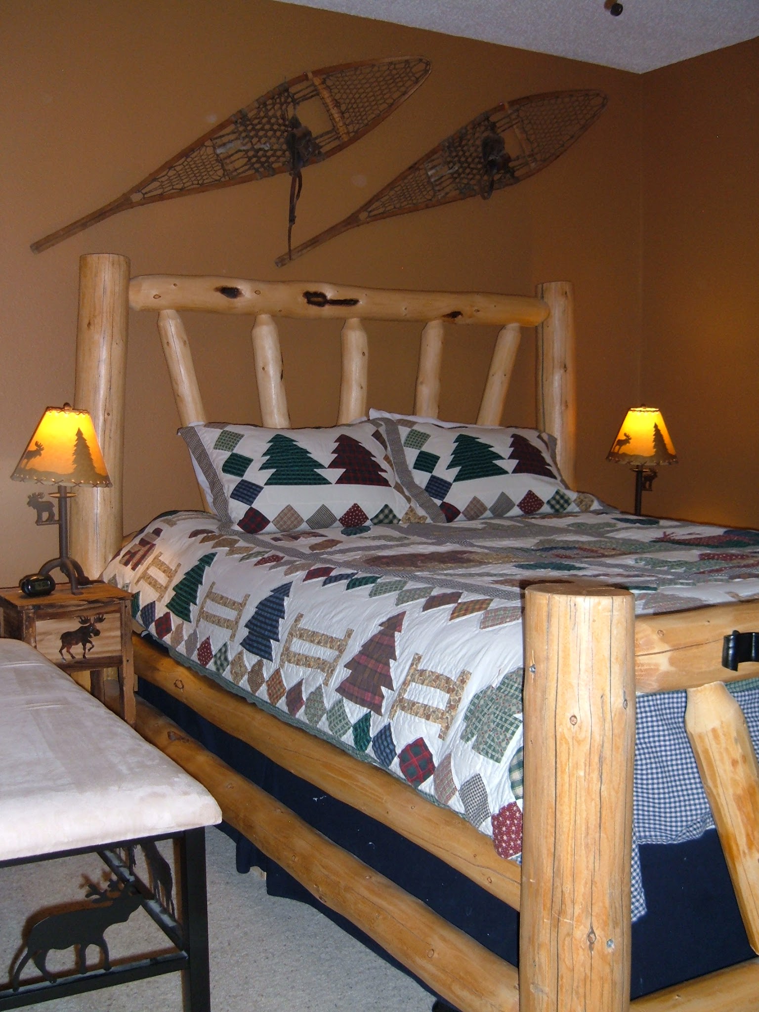 Bedroom with a log bed, down comforter, brown walls, snowshoes hanging on the wall, with beige carpet.  Moose theme bedside lamps and tables.