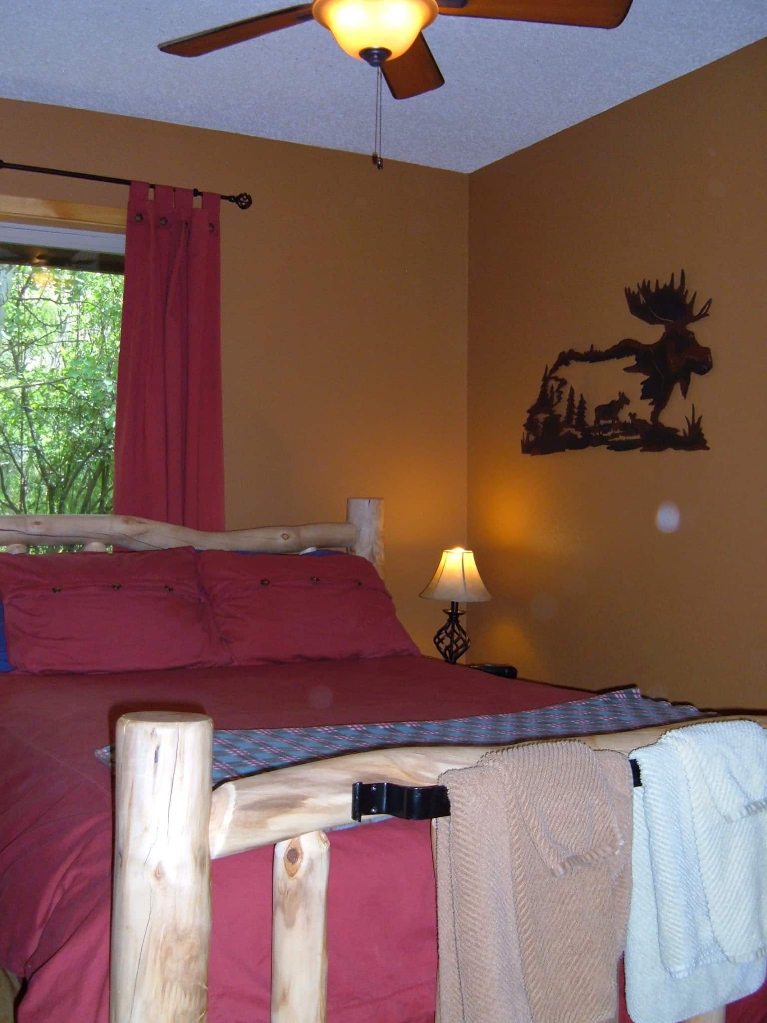 Charming log bed with a red duvet comforter, brown walls, metal art work of a moose on the wall, bed lamp, and a ceiling fan. Brown and green towel sets hanging on a rod iron towel bar at the foot of the bed.
