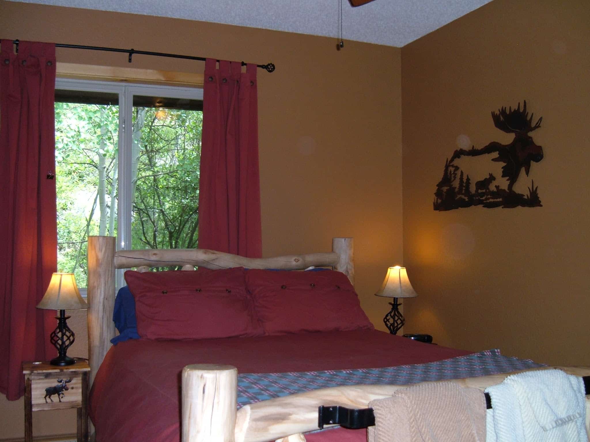Charming log bed with a red duvet comforter, brown walls, metal art work of a moose on the wall, bed lamp, and a ceiling fan. Brown and green towel sets hanging on a rod iron towel bar at the foot of the bed.