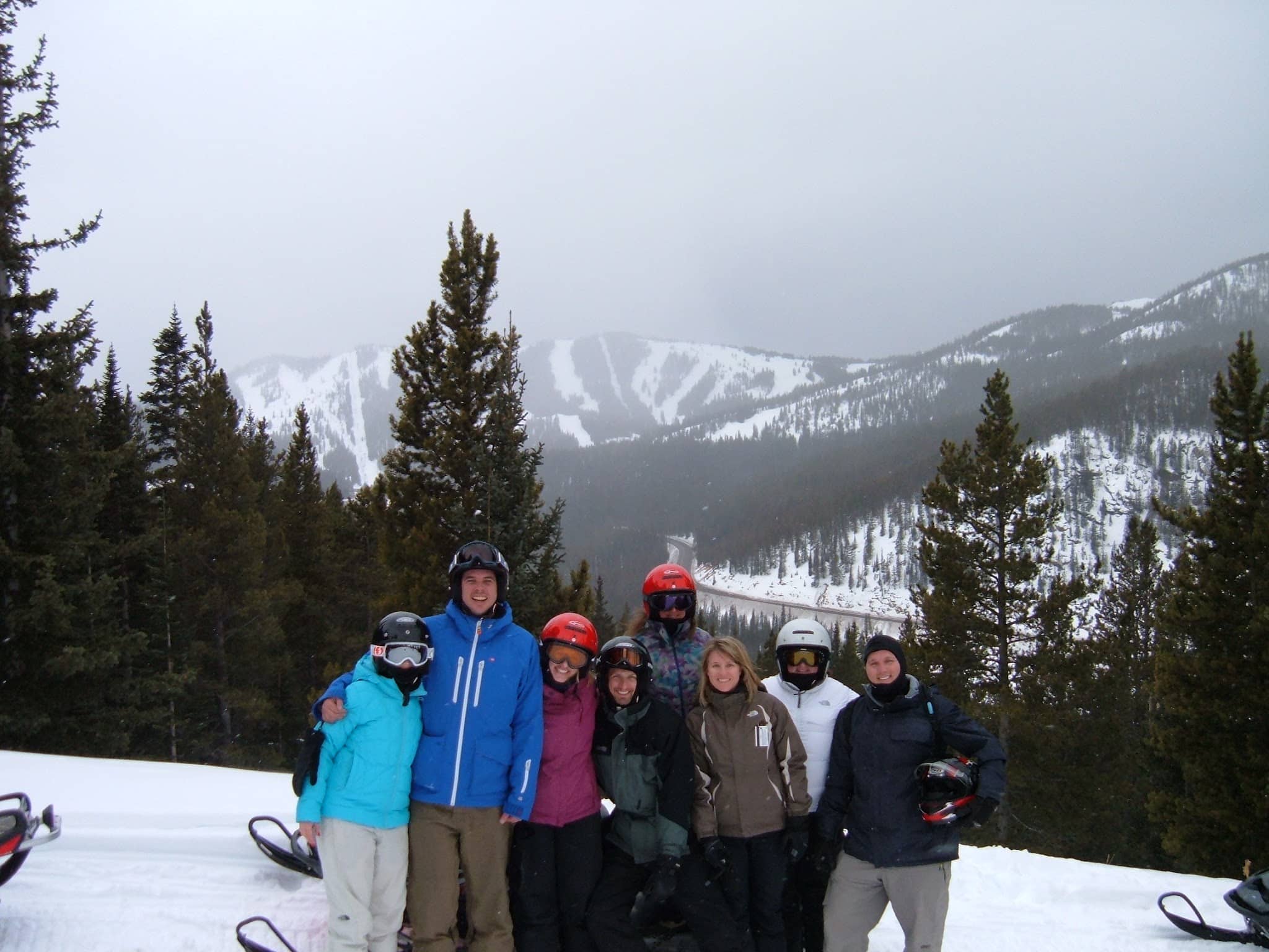 Eight people on a winter snowmobile tour with a ski area in the background.