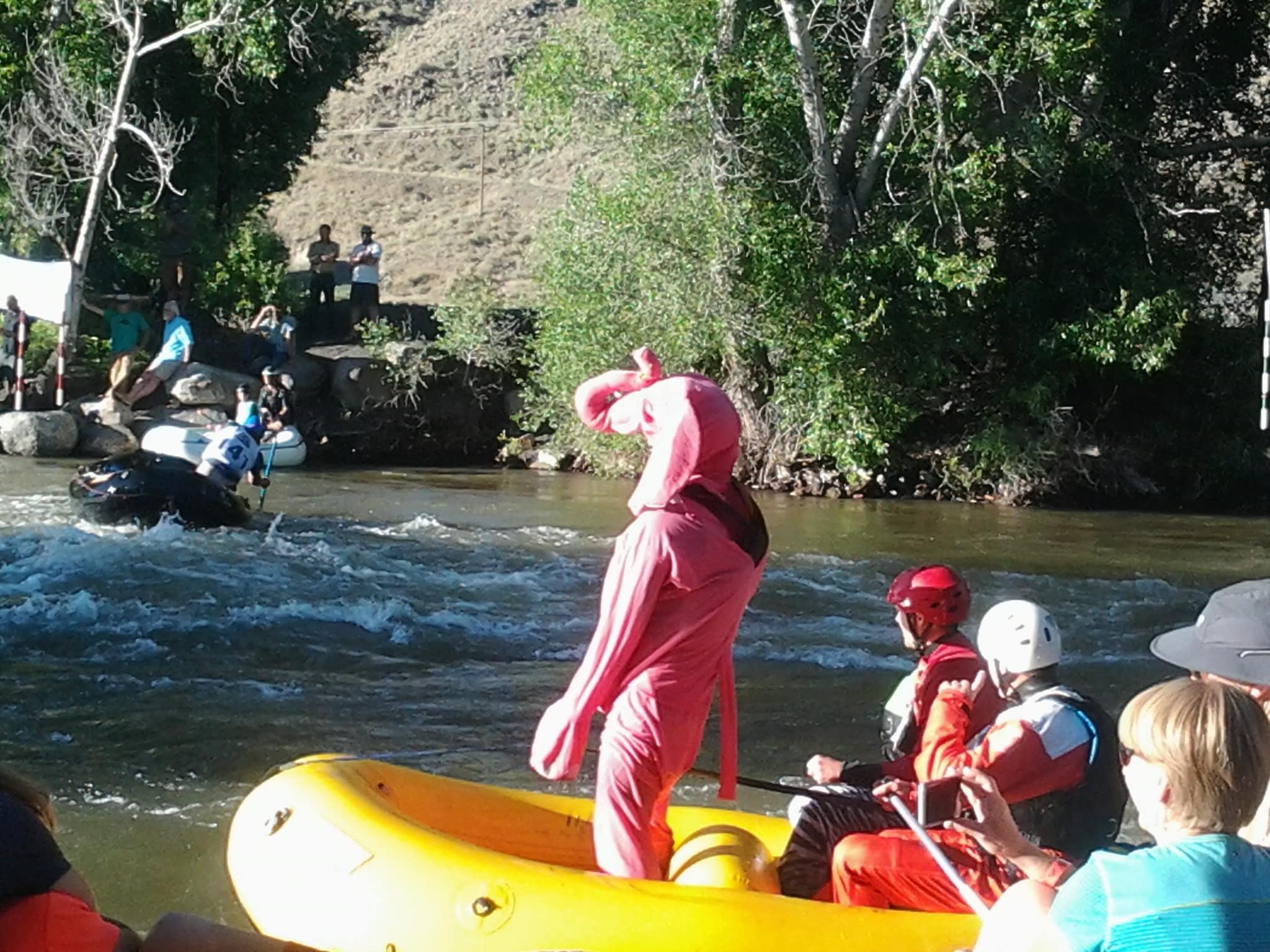 River competition with a person dressed in a pink elephant standing in the raft watching the other rafts, spectator all around.