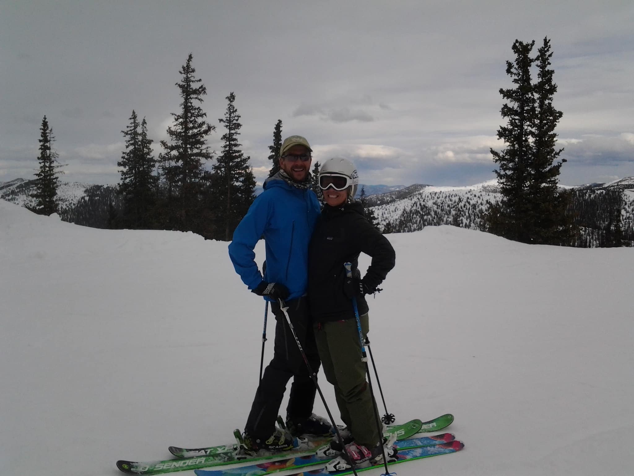 A couple skiing, posing for the camera with trees and mountains in the background.