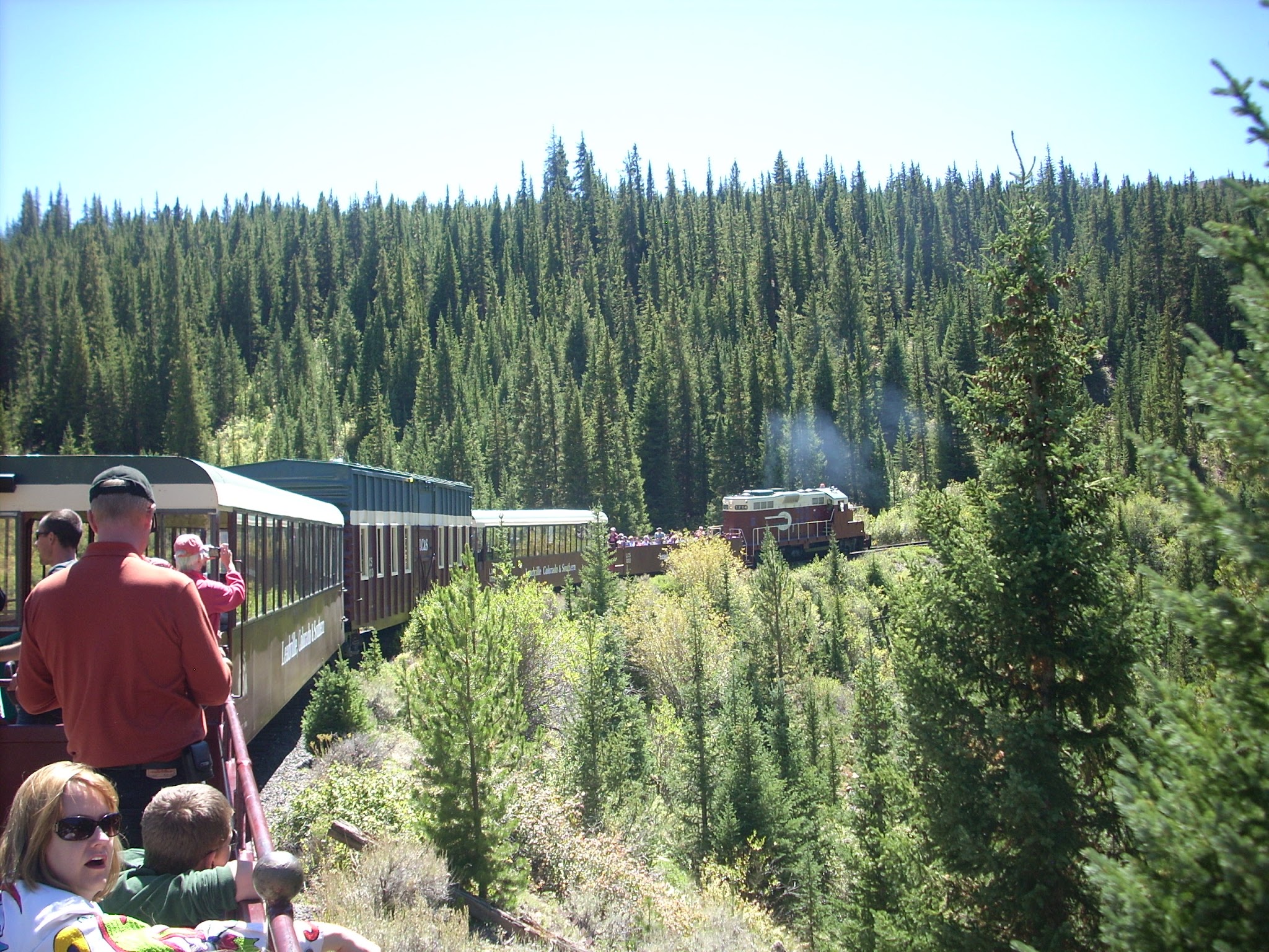 Passenger train ride along a mountain curve, with passengers taking photos and kids look out at the forest.