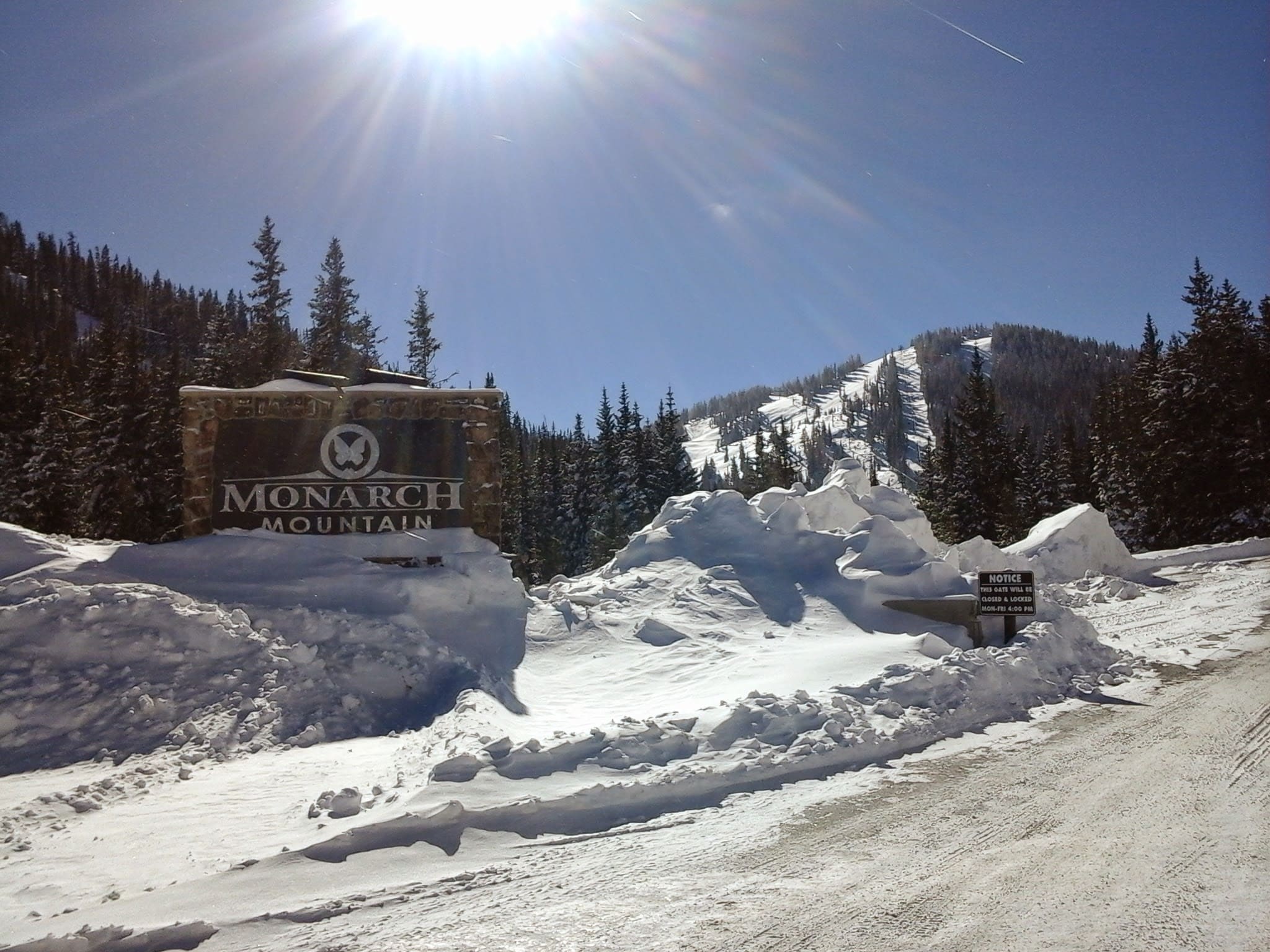 Sun shinning above the Monarch Mountain entrance sign, with the ski area in the distance all covered in snow.