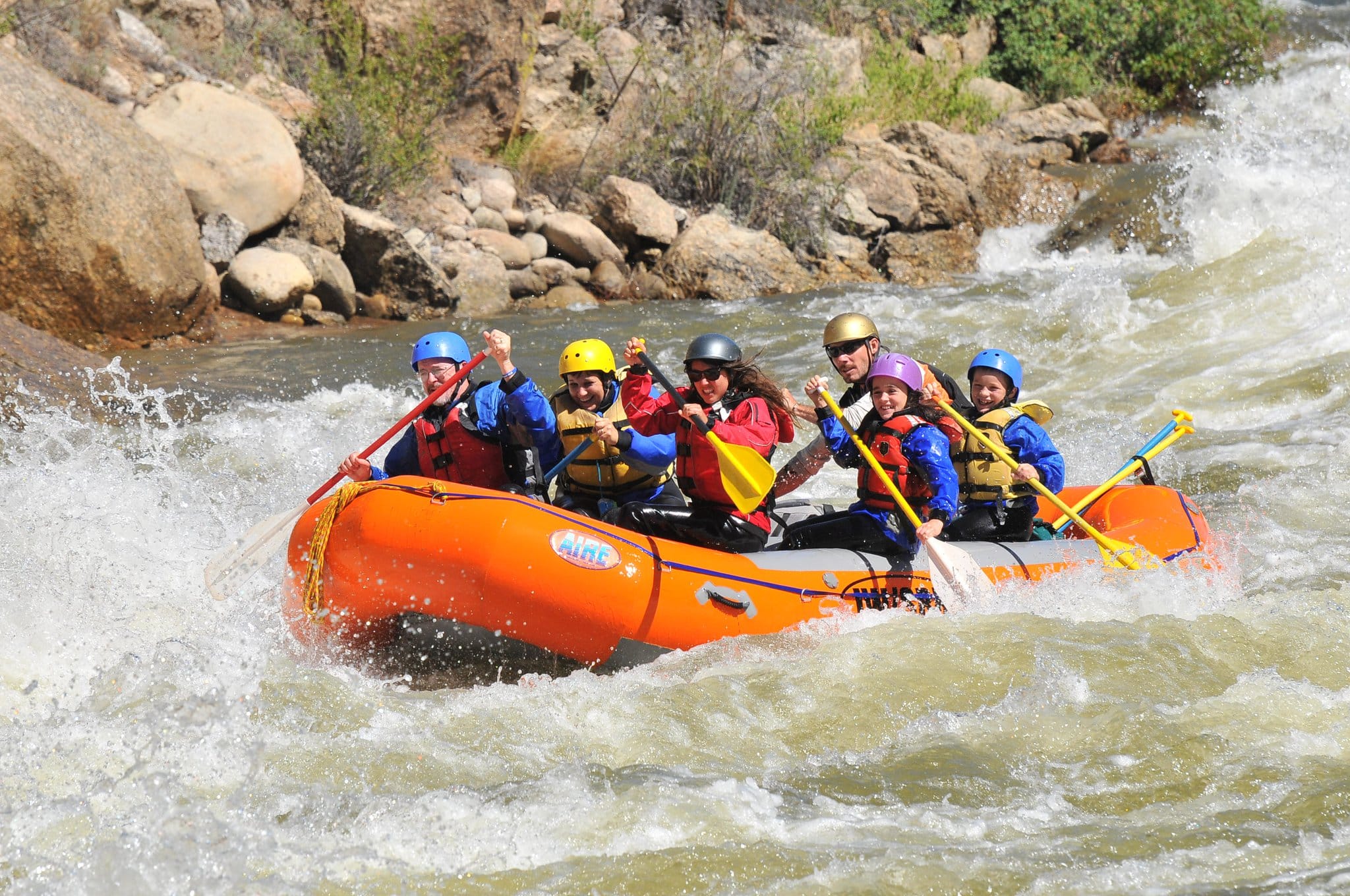 A raft filled with six guests paddling down a river in rapids on a sunny day.