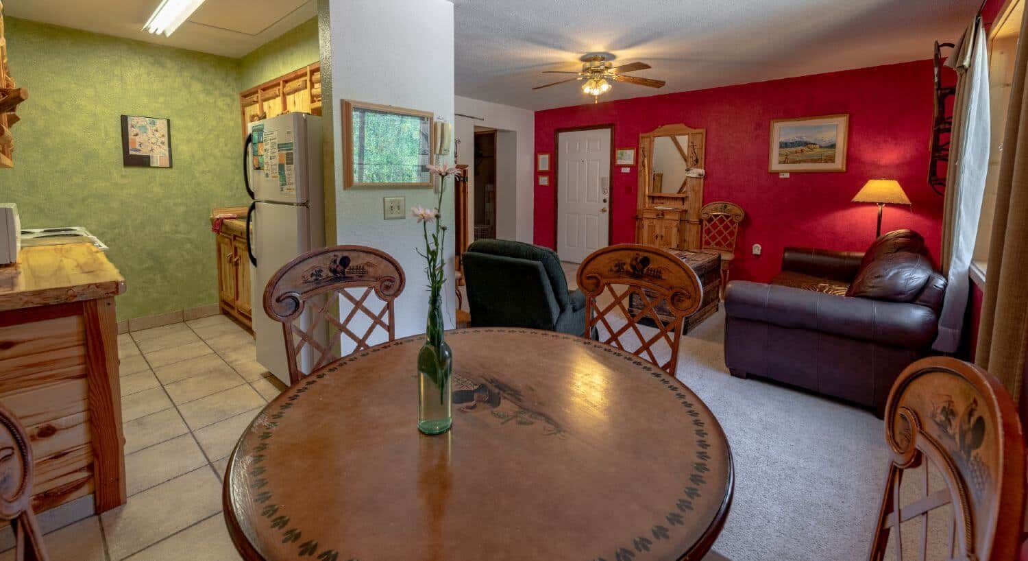 Dining table and 2 chairs, with kitchen to the left and living room with brown leather sofa, light carpet, red accent wall. TV rustic armoire. 