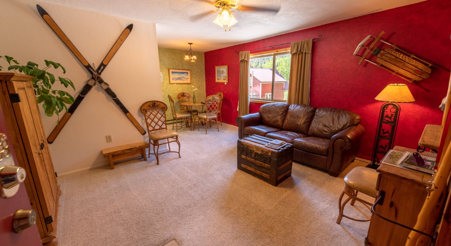 Condo B  living room with brown leather sofa, antique trunk as coffee table, antique skis crossed hanging on the wall, with light color carpeting; with dining table and 4 chairs back to the left. 