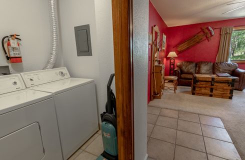 On the left, laundry room with white walls, on the right, view into the living room with red walls, carpeting, and brown leather sofa