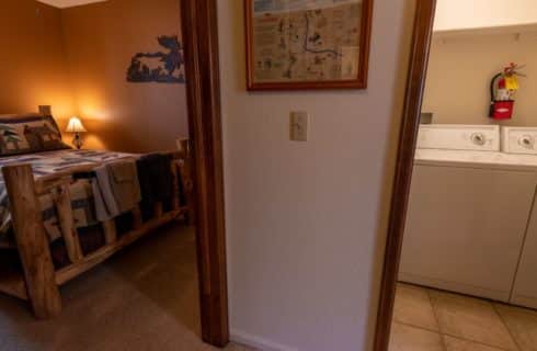 On the left, view into a bedroom with brown walls, carpeting, and wooden log bed, on the right, view into a laundry room with white walls and light colored tile flooring