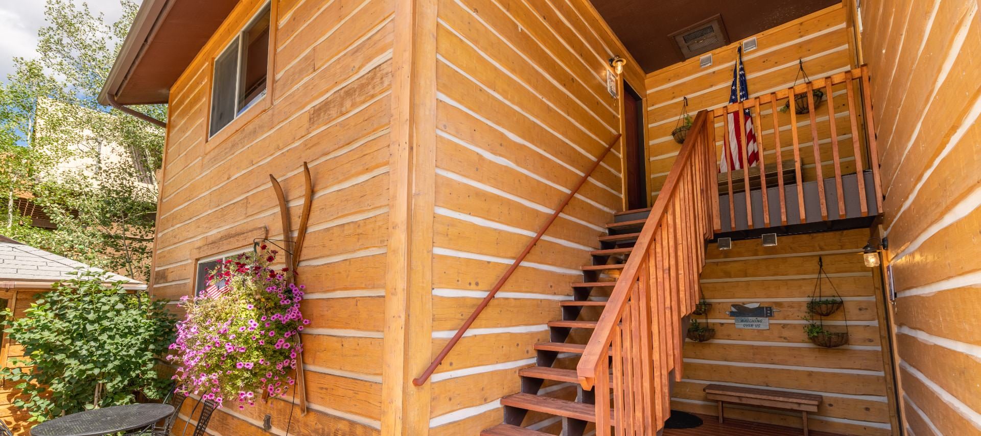 Exterior view of the property with wooden siding and staircase to second level surrounded by green trees and shrubs and flowers