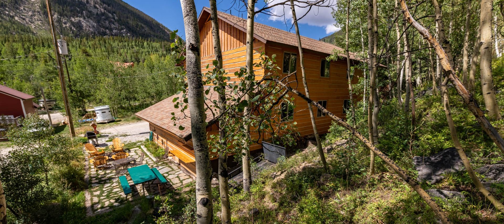 Exterior view of the property with wooden siding surrounded by green trees, shrubs and flowers, and a mountain