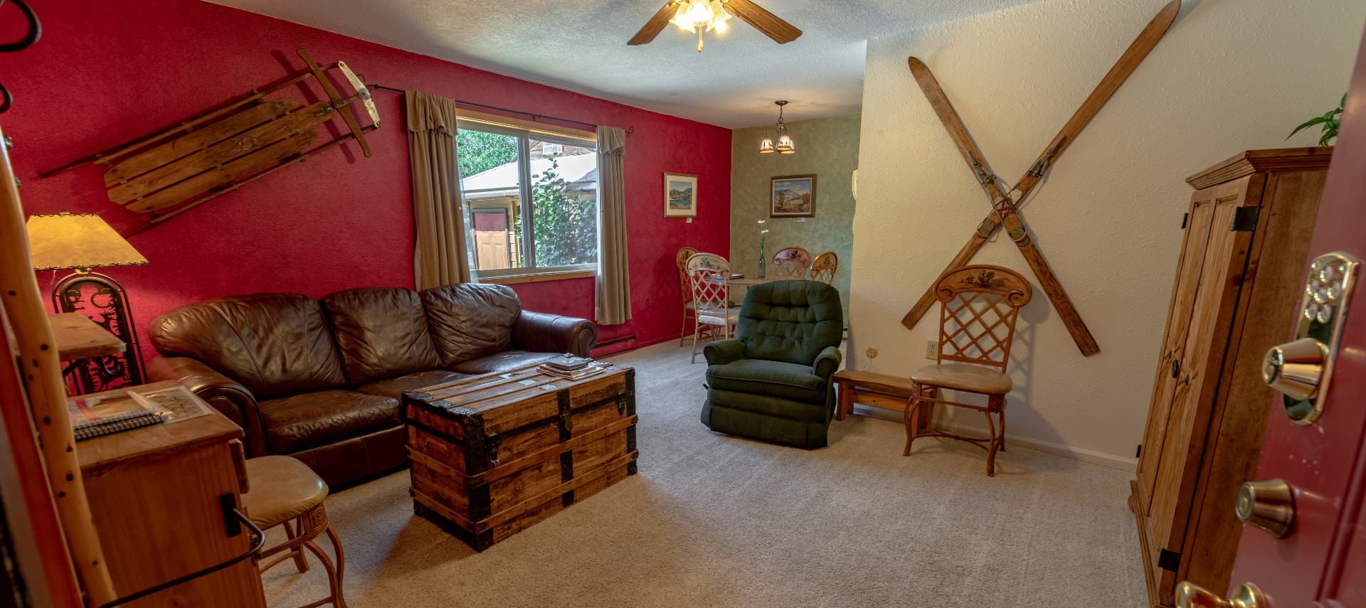 Living room with red, green, and cream walls, carpeting, brown leather couch, green arm chair, wooden trunk, and wooden hutch