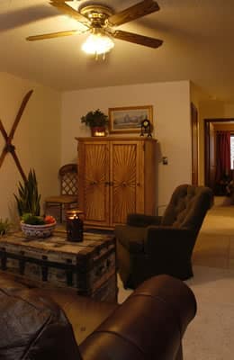 Living room with white walls, a brown leather sofa, chair, antique trunk as a coffee table, rustic armoire, wooden skis hanging on the wall, painting, and a ceiling fan.