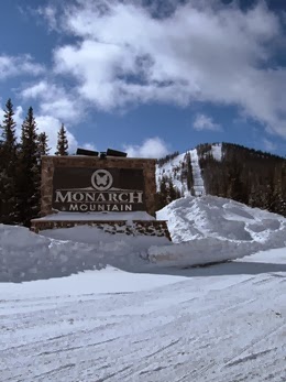 Sun shinning above the Monarch Mountain entrance sign, with the ski area in the distance all covered in snow.