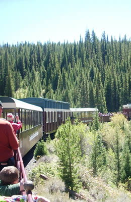 Passenger train ride on a big mountain curve on a sunny afternoon.  Passengers standing along the railing looking at the scenery and taking photos.