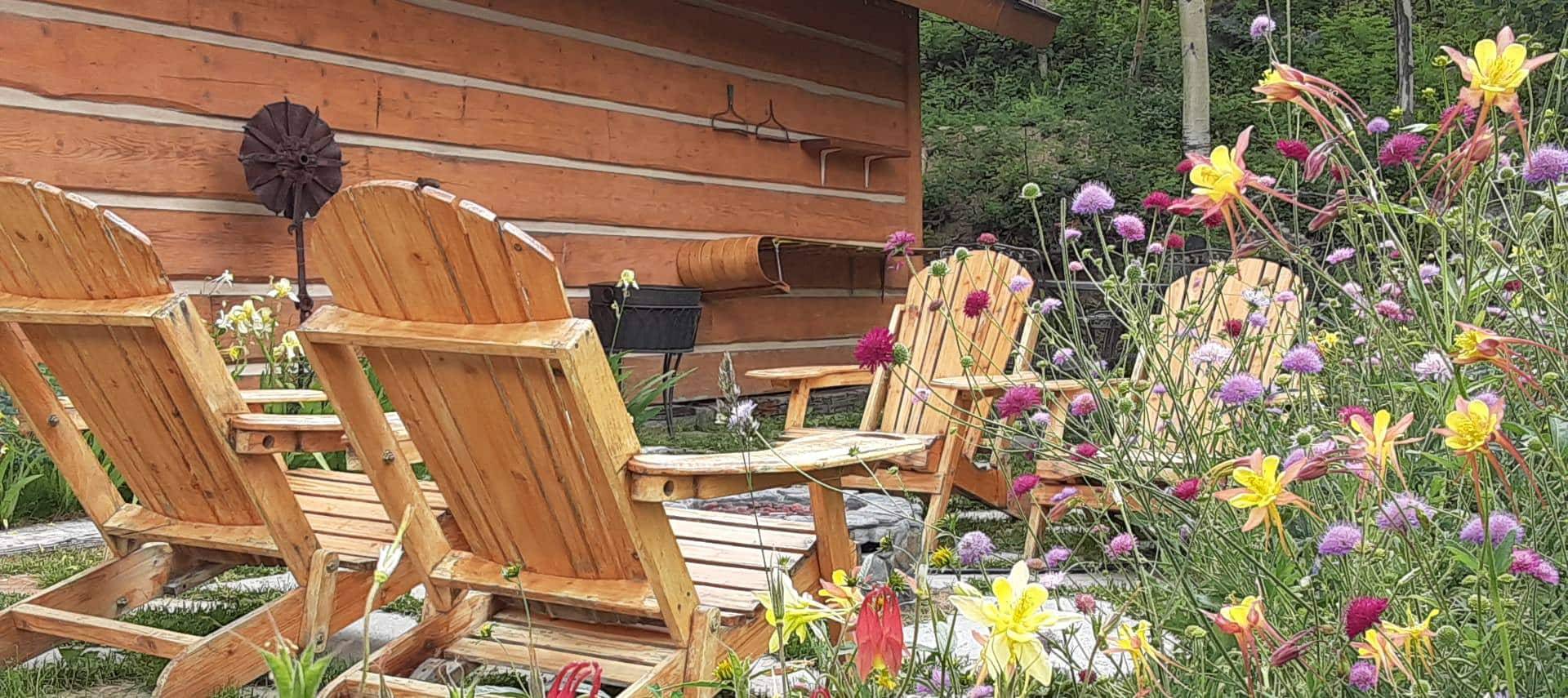 Four wooden adirondack chairs and a stone firepit surrounded by green grass and yellow, purple, and pink flowers