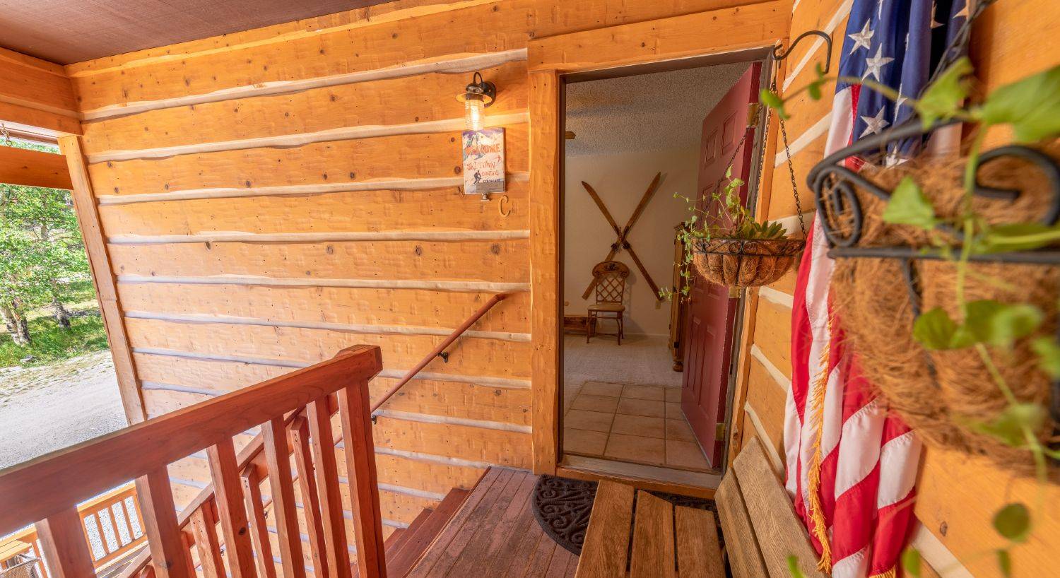 Entrance to condo C, hanging plants on the log siding, American flag standing in a holder, stairs leading down on the left, door open to the living room with antique skis on the wall.